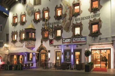 Spätgotisches Portal Ein traditionelles Hotelgebäude mit bunten Fenstern und schöner Fassade. Im Vordergrund ist ein Casino und Außensitzplätze sichtbar.
