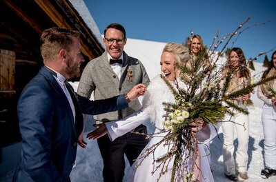 Hochzeitsplaner Finest Wedding Eine fröhliche Hochzeitsfeier im Schnee mit lächelnden Gästen. Die Braut trägt einen schönen Blumenstrauß und wird herzlich begrüßt.