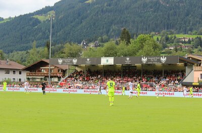 Ein Fußballspiel auf einem Stadionfeld mit Zuschauern im Hintergrund. Die umliegende Landschaft ist grün und bergig.