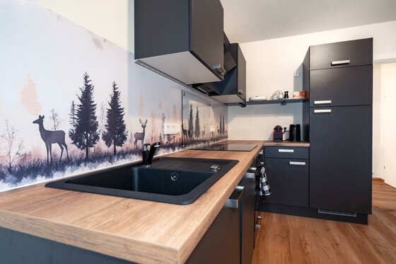 Kitzbühel_DeNi Apt_Küchenschrank Large Eine moderne Küche mit dunklen Schränken und einer Holzarbeitsplatte. Die Wand ist mit einem dekorativen Motiv von Rehen und Bäumen verziert.