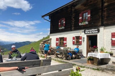 Gäste auf der Terrasse des Alpengasthauses Wiegalm vor einer Holzfassade mit Bergen im Hintergrund.