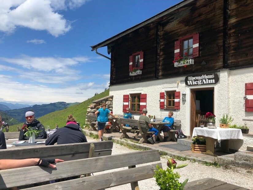 Terrasse der Wiegalm Gäste auf der Terrasse des Alpengasthauses Wiegalm vor einer Holzfassade mit Bergen im Hintergrund.