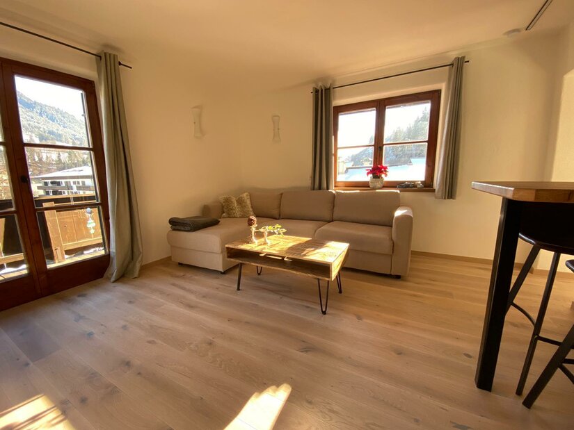 Wohnbereich mit beigem Ecksofa, Holztisch und Parkettboden vor einem Fenster.
