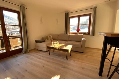 Wohnbereich mit beigem Ecksofa, Holztisch und Parkettboden vor einem Fenster.