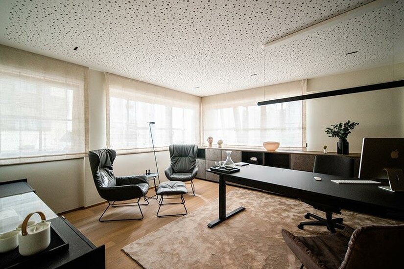 Ordinationsraum in Kitzbühel Modernes Büro mit Schreibtisch, zwei Ledersesseln und einer perforierten Decke.