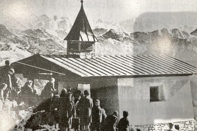 Maria Heimsuchung Ein historisches Bild einer kleinen Kapelle in den Bergen. Menschen stehen um das Gebäude herum, während schneebedeckte Gipfel im Hintergrund sichtbar sind.