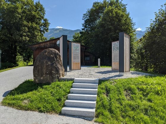 Aurach Wander-Infoplatz Infoplatz mit Steinstufen, Informationstafeln aus Holz und Metall sowie einem großen Findling vor Bäumen.