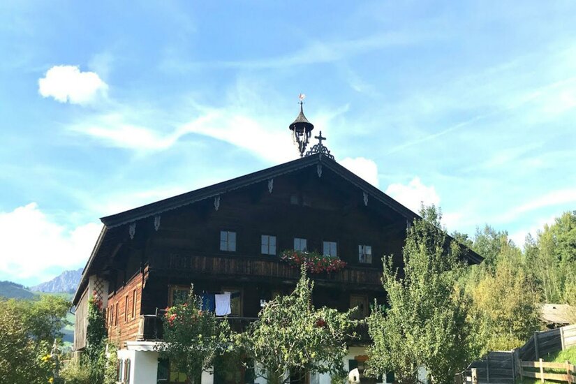 Ein traditionelles Holzhaus mit einem spitzen Dach und einem kleinen Turm. Umgeben von Bäumen und einem klaren Himmel.