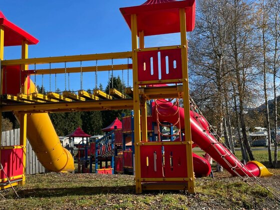 Bunter Spielplatz mit Klettertürmen, Hängebrücke und Rutschen vor Bäumen unter blauem Himmel.