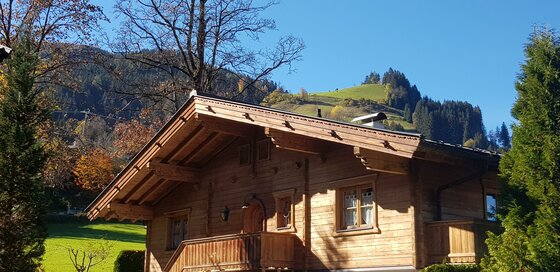 Blick auf Happy holiday Kitz Alpines Holzhaus mit Balkon vor bewaldeten Bergen unter blauem Himmel.