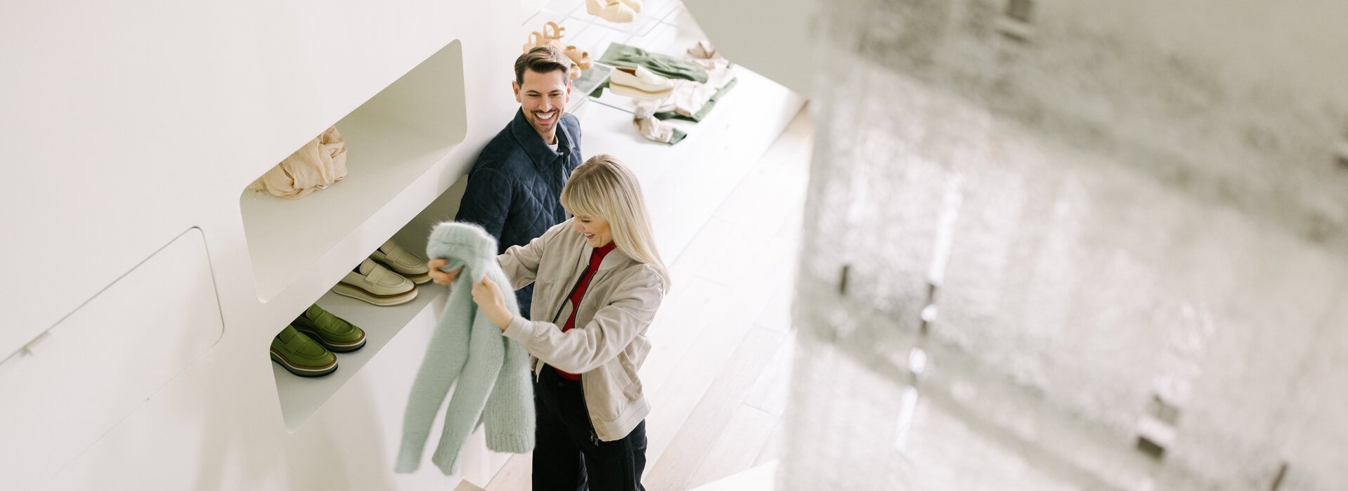 A young couple shopping in a bright store, the blonde woman is holding a light blue wool coat in her hands and smiling