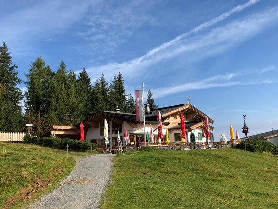 Berghütte mit Terrasse, Sonnenschirmen und Fahnenmast vor einem Nadelwald unter blauem Himmel.