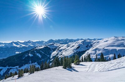 Verschneite Skipiste im Vordergrund mit Blick auf bewaldete Hänge und alpine Berggipfel unter blauem Himmel mit Sonne.