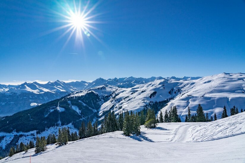 Verschneite Skipiste im Vordergrund mit Blick auf bewaldete Hänge und alpine Berggipfel unter blauem Himmel mit Sonne.