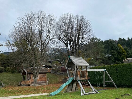 Kinderspielplatz aus Holz mit Rutsche, Klettergerüst und Schaukel vor Bäumen.