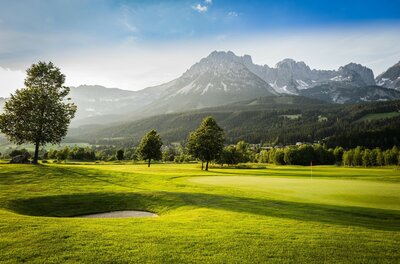 Ellmau_Golfschlaeger_Wilder Kaiser Eine malerische Golfanlage mit sattem Grün und sanften Hügeln. Im Hintergrund erheben sich majestätische Berge unter einem klaren Himmel.