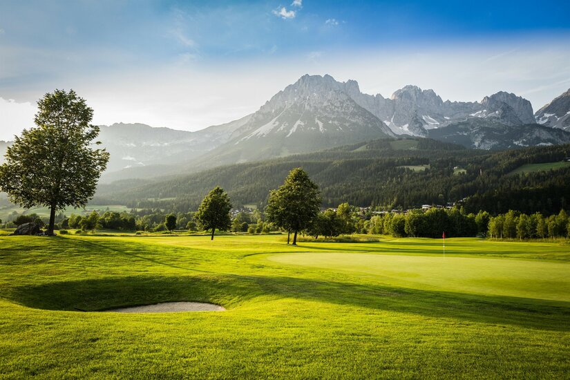 Eine malerische Golfanlage mit sattem Grün und sanften Hügeln. Im Hintergrund erheben sich majestätische Berge unter einem klaren Himmel.