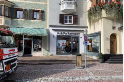 Behindertenparkplatz - Bichlstraße 8 Ein Einkaufsgeschäft mit dem Namen "Peak Performance" ist sichtbar. Daneben befinden sich weitere Läden und eine gepflasterte Straße.