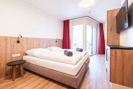 KitzSki Living Apartment Schlafzimmer Modernes Doppelzimmer mit hellem Holz, weißen Betten, roten Vorhängen und Balkonzugang.