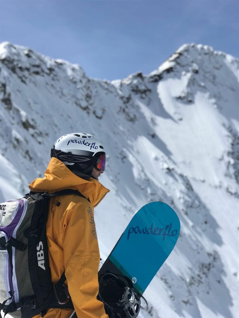 Snowboarder vor Berggipfel Snowboarder mit gelber Jacke, Helm und Rucksack blickt auf einen verschneiten Berggipfel.