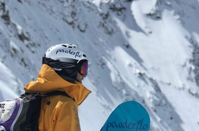 Snowboarder mit gelber Jacke, Helm und Rucksack blickt auf einen verschneiten Berggipfel.