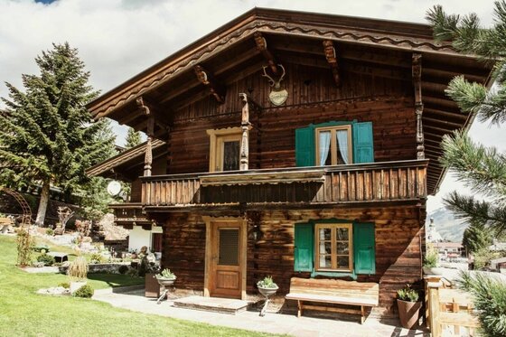 Zweistöckiges Holzhaus mit Balkon, grünen Fensterläden und einer Holzbank davor im Garten.