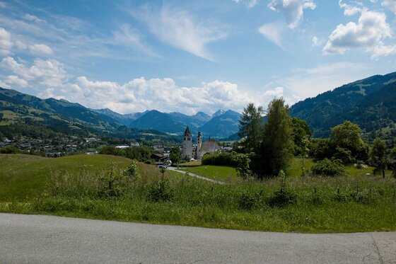 Dorfansicht Achrain mit Bergpanorama Blick über grüne Wiesen auf ein Dorf mit Kirchtürmen vor einer alpinen Bergkulisse unter blauem Himmel.