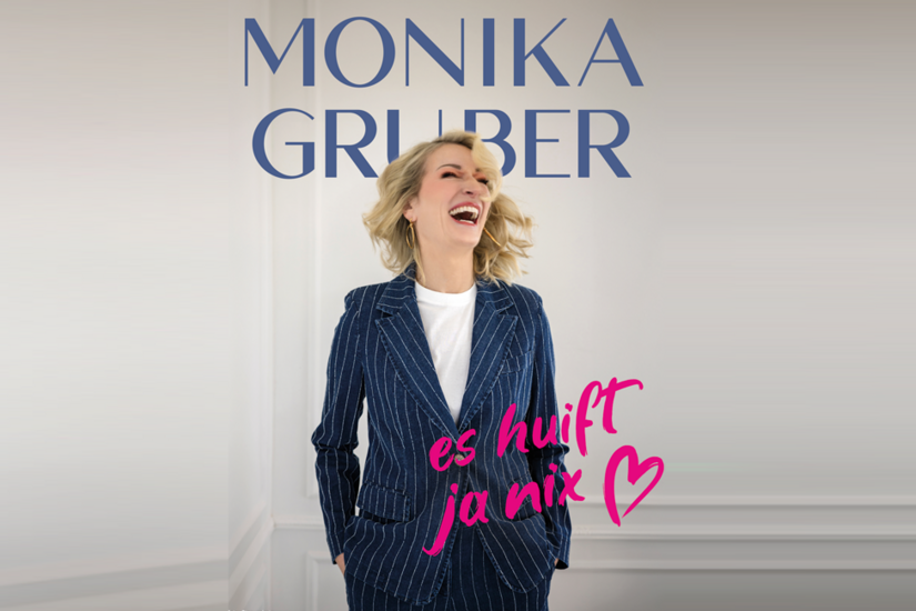 Porträt von Monika Gruber in einem blau-weiß gestreiften Anzug vor hellem Hintergrund.