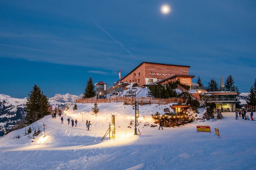 Beleuchtetes Berggasthaus am Hahnenkamm bei Nacht mit verschneitem Vorplatz und Spaziergängern.