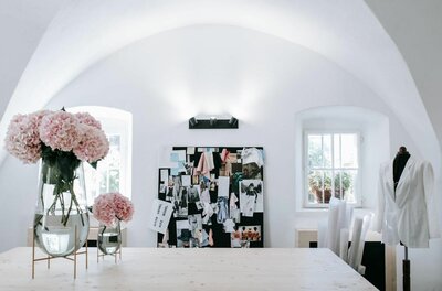 Heller Atelierraum mit Holztisch, rosa Hortensien in Glasvasen, Moodboard und Schneiderpuppe unter einem Gewölbe.