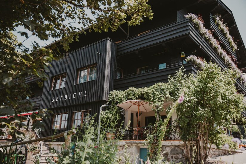 Hotel Seebichl im Sommer Außenansicht des Hotels Seebichl mit dunkler Holzfassade, Balkonen und einer begrünten Terrasse mit Sonnenschirm.
