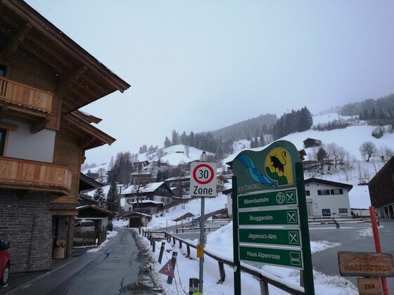 Verschneite Dorfstraße mit Hinweisschildern zu Almen und Bergbahnen vor bewaldetem Hang.
