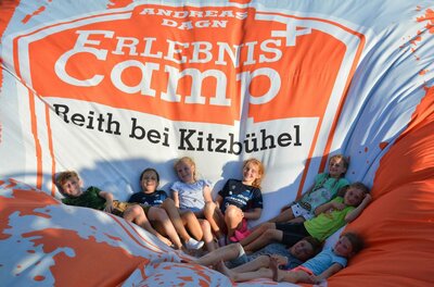 Erlebniscamp Reith bei Kitzbühel Kinder sitzen gemeinsam auf einem großen, orange-weißen Luftkissen mit der Aufschrift Erlebniscamp.