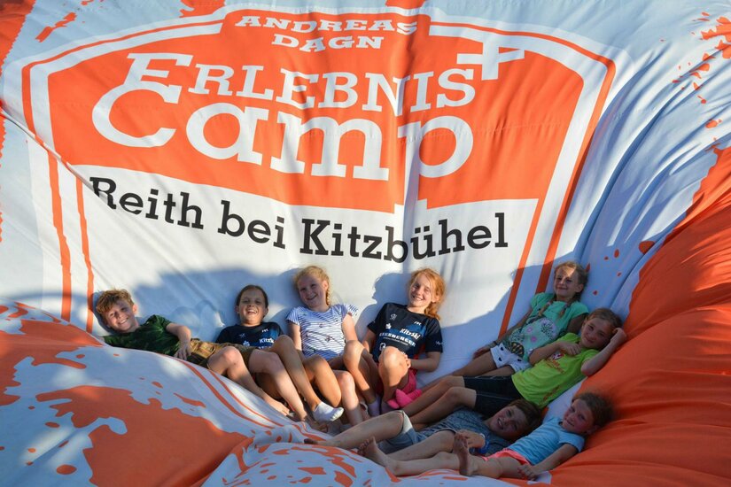 Erlebniscamp Reith bei Kitzbühel Kinder sitzen gemeinsam auf einem großen, orange-weißen Luftkissen mit der Aufschrift Erlebniscamp.