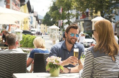 Ein Mann und eine Frau sitzen an einem Tisch im Außenbereich eines Cafés in einer belebten Fußgängerzone.