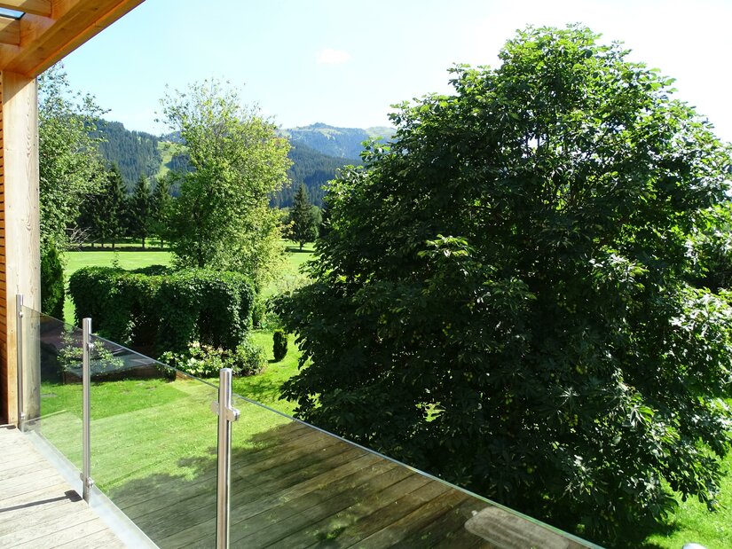 Balkonausblick am Golfweg Blick von einem Holzbalkon mit Glasgeländer auf eine grüne Gartenanlage mit Bäumen und Bergen im Hintergrund.