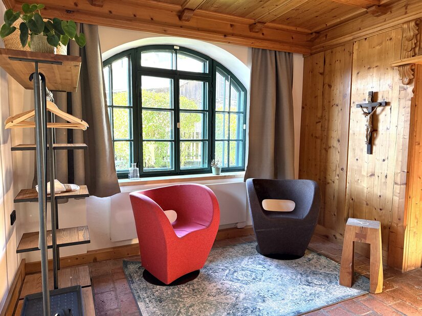 Ein gemütlicher Raum mit zwei modernen Sesseln in Rot und Schwarz. Ein großes Fenster sorgt für viel Licht und ein Holzregal ergänzt die stilvolle Einrichtung.