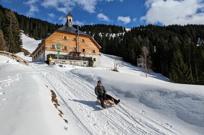 Rodeln bei der Kelchalm Person fährt auf einem Holzschlitten eine verschneite Bahn vor der Kelchalm hinunter.