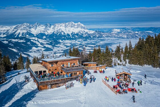 Berghaus Tirol im Winter Berghütte aus Holz mit Sonnenterrasse in einem verschneiten Skigebiet vor markantem Gebirgspanorama.