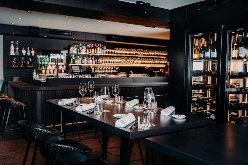 Les Deux Brasserie & Bar Gedeckter dunkler Holztisch mit Weingläsern und Servietten vor einer Bar mit beleuchtetem Weinregal.