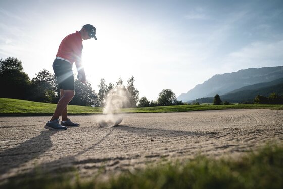 Ein Golfer schlägt einen Ball aus dem Sandbunker. Im Hintergrund sind Bäume und Berge zu sehen.
