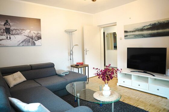 2916768_960h Ein gemütliches Wohnzimmer mit einer grauen Couch und einem Glastisch. An der Wand hängen Bilder von Berglandschaften, und ein Fernseher steht auf einem weißen TV-Tisch.
