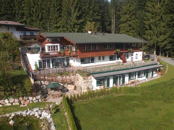 Mehrstöckiges alpines Holzhaus mit weißem Erdgeschoss vor einem Nadelwald.
