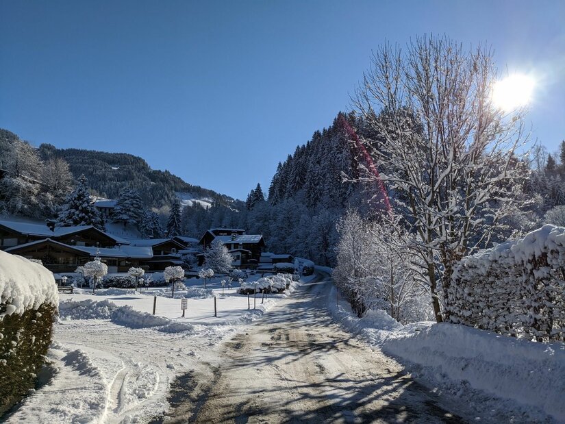 Winterliche Siedlung Oberaigen Verschneite Straße in einer alpinen Siedlung mit Holzhäusern und bewaldeten Bergen unter blauem Himmel.