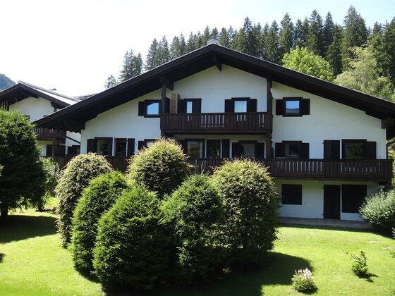 Außenansicht Haus Fleck Zweistöckiges weißes Haus mit dunklen Holzbalkonen und grünen Sträuchern im Vordergrund.
