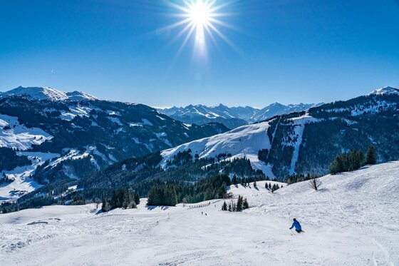 Ein Skifahrer auf einer präparierten Piste vor verschneiten Berggipfeln unter strahlender Sonne.