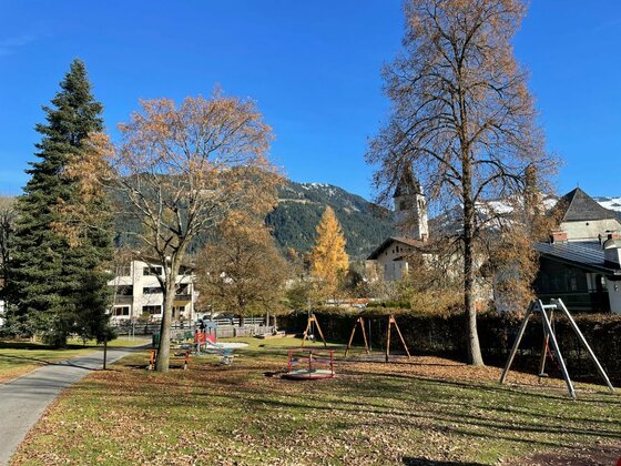Ein öffentlicher Spielplatz mit Schaukeln und Karussell vor einer Bergkulisse im Herbst.