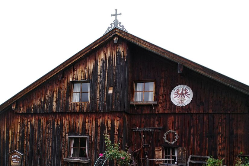 Eine rustikale Holzhütte mit einem Kreuz auf dem Dach. An den Wänden sind Fenster und ein Wappensymbol zu sehen.