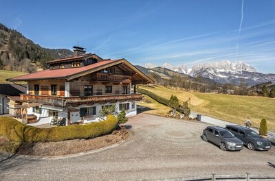 Außenansicht mit Wilder Kaiser Alpines Holzhaus mit Balkon vor einer Bergkette unter blauem Himmel.