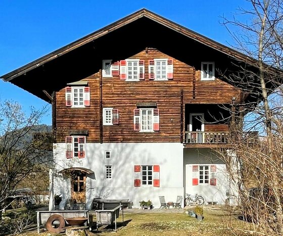Ein traditionelles Holzhaus mit roten Fensterläden und einem großen Dach. Es liegt in einer ländlichen Umgebung mit Bäumen und einem klaren Himmel.